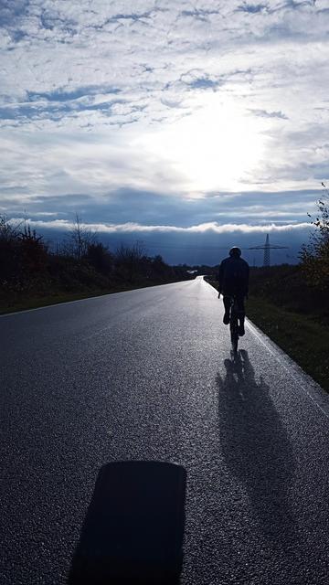 Ein Rennradfahrer auf einer Landstraße. Es ist bewölkt und die durch die Wolken brechende Sonne spiegelt sich sanft auf dem naßem Asphalt.