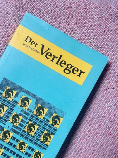 Ein Foto des Buchcovers »Der Verleger« auf grobem roten Stoff. Das Buch liegt schräg, ist unten angeschnitten . Auf grünlichem Grund (falsche Farbdarstellung auf dem Foto) oben ein gelber Balken, darin Titel und Autor. Unten mittig auf dem Cover eine Montage eines schwarz-weiß Bildes, eingefärbt im Ton des Covers. Auf dem Bild ein Hochhaus, darauf mehrere identische, symetrisch angeordnete Portraits (s/w, gelb eingefärbt) einer Person, welche in die Kamera schaut, eine Hand an der Stirn vielleicht um besser sehen zu können oder Sonnenlicht abzuhalten.