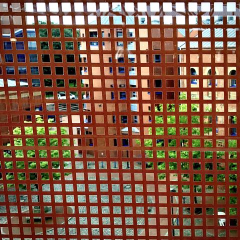 The photo shows a view of the Bremerhaven University of Applied Sciences campus, taken from the third floor of Building Z. The scene is photographed through the building’s heat- and sun-protection screen, which creates a grid of small square openings across the entire image. Through the grid, you can see the courtyard area leading toward the roundabout (“Rondell”) and Building K. Red-brick façades, blue window frames, patches of greenery, and the paved courtyard are visible, slightly abstracted by the patterned screen in the foreground.