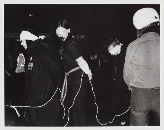 The image depicts a group of individuals participating in what appears to be an anti-Vietnam war demonstration, as indicated by the caption. It is a black and white photograph showing four people standing closely together at night.

Two persons on the left are dressed in nun-like habits with their heads covered by hoods or bonnets that resemble those worn by religious nuns, suggesting they may be portraying Catholic clergy members. One of them holds onto what appears to be an extension cord loosely draped over her body and floor, which is connected to a device not visible in the frame.

In the center stands another person wearing dark clothing with their face partially obscured by white material that could represent a mask or blindfold, holding similar cords as those on either side. This figure's attire seems more casual compared to the nun-like costumes of the others.

To the right is an individual dressed casually in what looks like jeans and a leather jacket; they wear a motorcycle helmet for protection, which contrasts with the religious themes carried by their companions.

The fourth person stands slightly apart from the group on the far right. They are wearing more casual attire consisting of grey pants and a light-colored jacket or coat without any visible headgear that matches the others' outfits.

In summary, this image captures an intense moment during a protest march with people in different attires expressing [...]