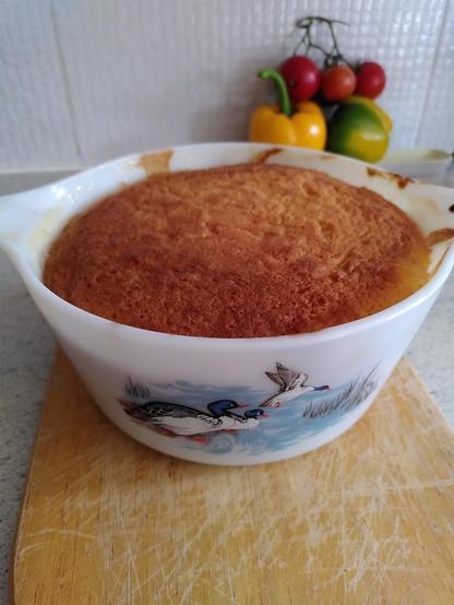 Pyrex dish with ducks on a pond  pattern on the front. It contains an apple and sponge pudding which is a golden brown. In the background a few homegrown tomatoes and peppers