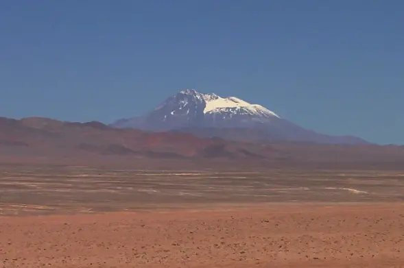 The image depicts a mountainous landscape with a prominent snow-capped peak in the background, set against a clear blue sky. The foreground features a dry, barren area with sparse vegetation, typical of arid environments. This type of scenery is often found in high-altitude regions or desert areas.

Image Credits: Wikimedia / Bachelot Pierre J-P / CC BY-SA 3.0