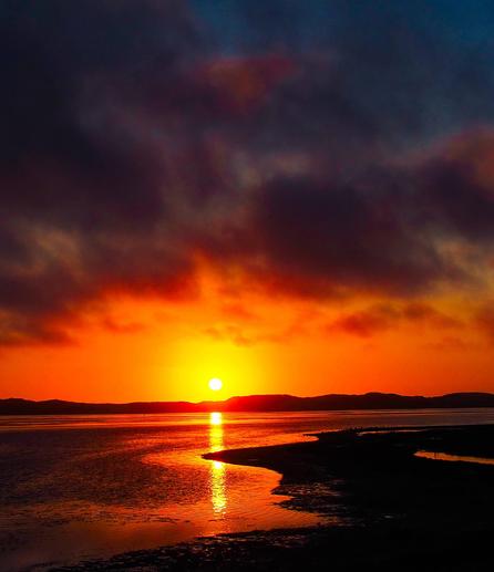 A vibrant, wide-angle landscape photograph depicts a sunset over a body of water. The sky is dominated by shades of orange and red, with a bright, circular sun positioned near the horizon. The water reflects the intense colors of the sky, creating a shimmering, golden path towards the viewer. A dark, silhouetted landmass stretches across the bottom of the frame, with a winding channel of water leading towards the sun. The image is mostly comprised of warm tones, with the landmass providing a stark contrast to the fiery sky and water.