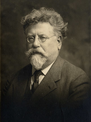 A sepia-toned portrait of an older man with wavy hair, a full mustache, and a pointed beard. He wears round glasses and a dark suit with a tie, looking slightly past the camera with a serious expression. The background is softly blurred.