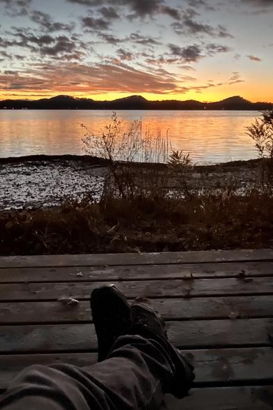 Your view over a lake at sunset. In the foreground you can see legs stretched out on an old, wooden deck with peeling stain and dead autumn leaves as you take in the view. At the edge of the deck are dried stems of reeds and aquatic plants. The lake reflects orange and yellow. On the horizon are sand dunes, and above them are yellow and orange clouds. Higher in the sky are dark clouds which have already slipped past the reach of the setting sun.