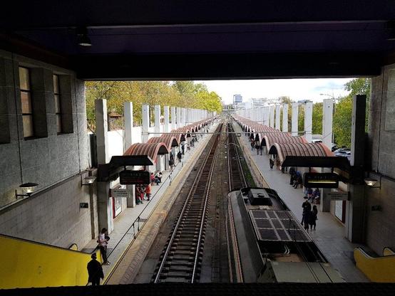 La estación de Delicias (Madrid). Dos hileras de bóvedas sobre los andenes (cada bóveda perpendicular al andén) y en medio las dos vías con traviesas metálicas sobre balasto de hormigón. A la izquierda, árboles altos que superan la altura de los tensores de las bóvedas. A la derecha, más bajos (la calle es más baja por ese lado). Al fondo, una torre de hormigón y cristal ligeramente a la derecha del punto de fuga. El cielo tiene nubes.