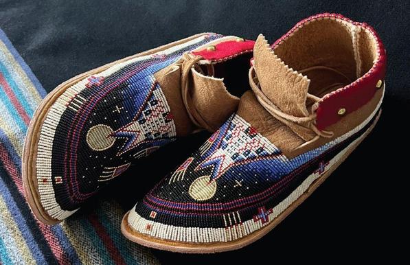 A pair of beaded moccasins. The design on the moccasins are of a tipi at night, where the sky is clear and the stars and a full moon shine down on everything. Off to the side of the tipi are what appears to be a pair of bear claws, reaching up towards the night sky.