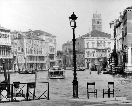 This black and white photograph captures a historic view of Venice's iconic Grand Canal. The perspective is from the edge of the canal, looking down its length with buildings on both sides that exhibit Venetian architectural style, characterized by their ornate facades adorned with balconies and intricate window designs.

The waterway is populated with various boats: a larger steamboat appears in motion at the center distance, while more distant to it are smaller gondolas typical of Venice. On either side of this canal view, traditional Venetian chairs sit empty on the banks, suggesting an atmosphere that has both historical significance and contemporary use.

A prominent street lamp with a classic design is centered foregrounding the scene. The sky above appears overcast or foggy, which softens the details of the buildings in the background but enhances their texture through contrast against this muted backdrop. There's no discernible human presence within the frame, adding to its timeless quality and quietude.

This image likely dates from a time before modern vehicles dominated Venice's streets as it prominently features such iconic elements that are still present today with a level of preservation suggesting an earlier era.