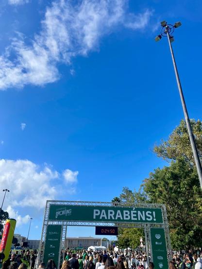 Runners crossing the finish line arch for the Corrida Sporting race. The banner reads "PARABÉNS!" (Congratulations!) and a digital clock displays the time 0:24:50.