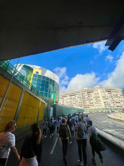 A group of runners on a road passing beneath an overpass, with the modern, yellow-sided exterior of the stadium complex visible on the left and a large, residential building in the background.