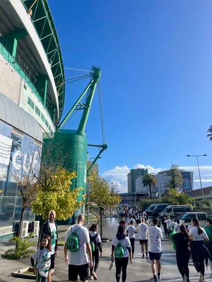 Runners in white and green shirts moving along a road outside the massive, green and white exterior of the Estádio José Alvalade (Sporting CP's stadium). The sky is clear blue.