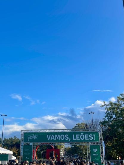 A large crowd gathered beneath the starting arch for the Corrida Sporting race. The banner reads "VAMOS, LEÕES!" (Go, Lions!) against a bright blue sky.