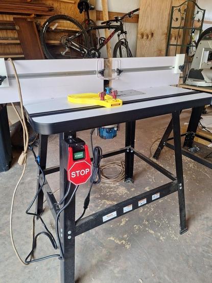 A fully set-up router table sits in a garage shop. The floor is lightly decorated with saw dust and wood shavings. The router table has a large On / Off switch installed, featuring an over-sized "Off" paddle, making it easy to turn the device off when needed. A full complement of safety features area also installed, including stop blocks, a dust hood and bit guard, and feather boards (only one is visible here, but I have additional boards to mount to the vertical guides when necessary. Routers are wonderful, but incredibly dangerous pieces of equipment, so I take safety seriously in the shop and especially around equipment such as this.