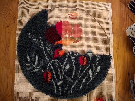 Auf einem Holzfußboden liegt ein etwa 60 Mal 60 cm großes Stück grobmaschiges Stramin, auf dem das Design für einen runden Teppich aufgedruckt ist. Das Design besteht aus einer großen roten Mohnblüte und vier Mohnblüten-Knospen, sowie Blätter, Ähren und Halmen in zwei Grautönen, das ganze auf einem fast schwarzen Hintergrund.
Der Teppich ist zu etwa zwei Dritteln fertig geknüpft.