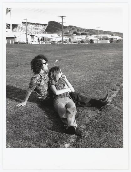 The photograph is a black and white candid shot featuring two individuals relaxing on grass. The adult appears to be wearing sunglasses, patterned short-sleeve shirt, trousers, and boots, with one hand resting on the ground for support while leaning against it casually. Beside them sits another individual in mid-motion of an arm gesture or reaching out, possibly a child judging by their smaller stature, dressed in what seems like a floral top paired with grey pants. In the background is open grassland leading up to buildings and structures that suggest this scene may be within a recreational field near urban surroundings. The sky appears clear suggesting fair weather conditions during the time of capture.