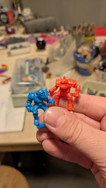 A hand holding two plastic miniatures. A blue human and a red orc. They are old hammer style 25mm chunky sculpts