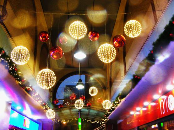 This photo shows a festive covered walkway decorated with glowing Christmas lights. Bright golden spheres made of tiny bulbs hang overhead, mixed with shiny red baubles that reflect the surroundings. Droplets on the lens soften the scene, creating hazy circles of light and giving the image a dreamy, atmospheric feel. Garlands with small red ornaments run along both sides of the corridor, while colourful shop fronts add blue and red tones to the lower edges. In the distance, through an archway, a faint glimpse of buildings is visible against the evening sky