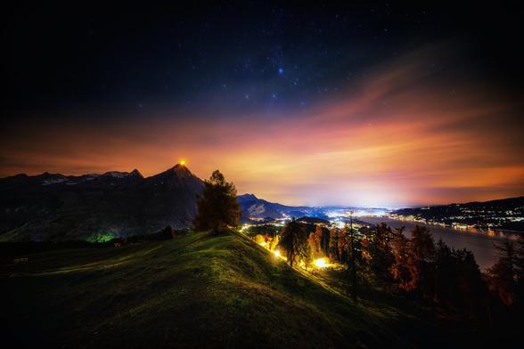 Eine stimmungsvolle Langzeitbelichtung bei Nacht zeigt einen weiten Blick über eine hügelige Landschaft mit dem Thunersee und der Stadt Thun im Hintergrund. Im linken Zentrum ragt der Berg Niesen hervor, dessen Spitze mit einem hell leuchtenden, goldgelben Lichtpunkt markiert ist. Das Himmelszelt darüber erstrahlt in einem tiefen Blau mit sichtbaren Sternen, während die Horizontlinie von einem intensiven Orange-Rot-Gelb des Lichtsmogs der dahinter liegenden städtischen Gebiete dominiert wird. Im Vordergrund erstreckt sich eine sanfte, dunkelgrüne Weidefläche, gekrönt von einem einzelnen Baum, dessen Äste sich silhouettenhaft gegen den leuchtenden Himmel abzeichnen. Rechts davon säumen dunkle Baumkronen den Hügelkamm. Die Ufer des Sees sind gesäumt von einer Kette warmer, weisser und gelber Lichter der Siedlungen.