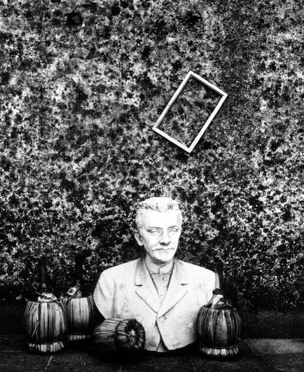 The black and white photograph depicts a bust of an elderly man with graying hair and a mustache, dressed in what appears to be clerical attire. The bust is centered on the table against a backdrop that resembles a weathered wall or textured surface. Surrounding the bust are various objects: two ornate vases resembling traditional Japanese lanterns with bamboo bases, one of which has its lid open revealing an interior; and several rolled-up newspapers or scrolls placed in front of them.

Above the sculpture is a rectangular picture frame tilted to the right side on the wall-like background. The image exudes a sense of antiquity, nostalgia, and possibly memorial significance due to the presence of the bust, which could be interpreted as depicting a figure of historical importance related to religion or academia.
