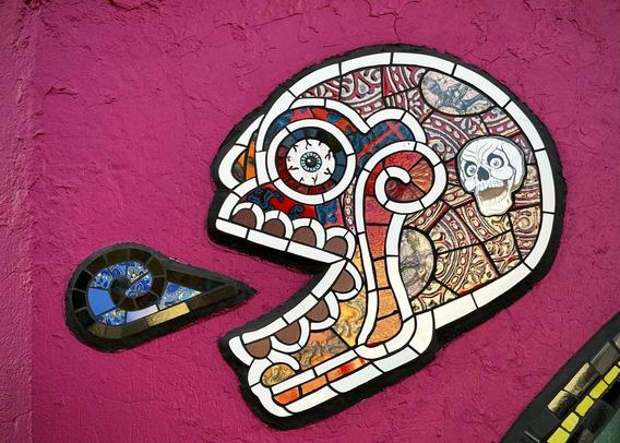 Urban mural detail of a colorful human skull speaking, made from reclaimed ceramic tiles on a magenta wall.