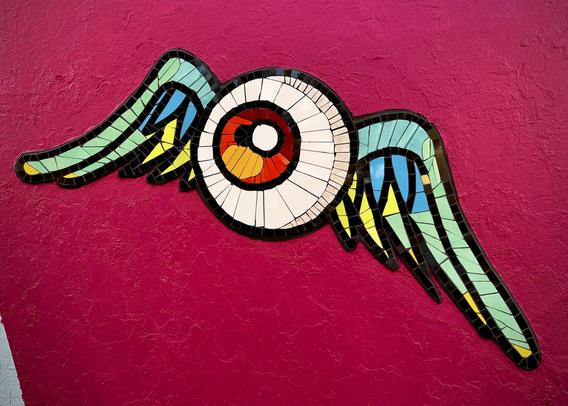 Urban mural detail showing an eyeball with an orange-red pupil and blue wings, made from reclaimed ceramic tiles on a magenta wall.