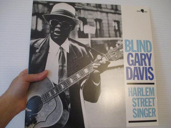 Réédition du 33T de Blind Gary Davis de 1960 "Harlem Street Singer", image en N&B du chanteur afro-américain et sa guitare en train de jouer dans la rue, chapeau blanc vissé sur la tête et lunettes noires rondes sur le nez. Un bâtiment de ville à l'arrière-plan