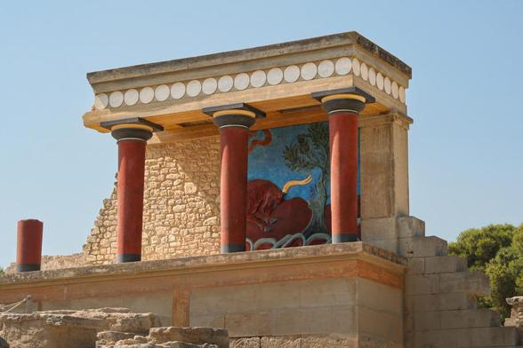 A partial view of a Minoan temple reconstruction featuring red columns, a colorful fresco of a bull, and stone walls under a clear blue sky.