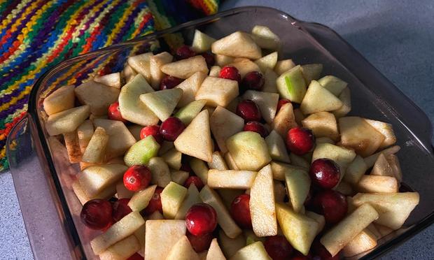 A square glass baking dish sitting on a kitchen counter with some colorful potholders nearby. The dish is filled with chunks of fresh-diced green and red apples, plus bright red cranberries, seasoned with brown sugar and cinnamon, waiting patiently for a crumbly topping and a bake in the oven.