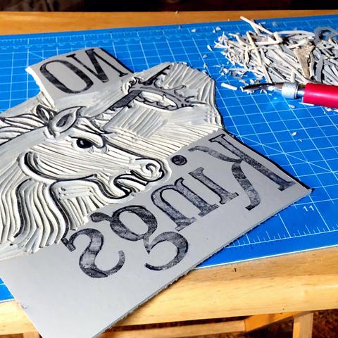 The same linoleum block from the post above but with a section of the black space around and inside of the unicorn carved away and a pink interchangeable lino cut tool next to a pile of linoleum shavings nearby