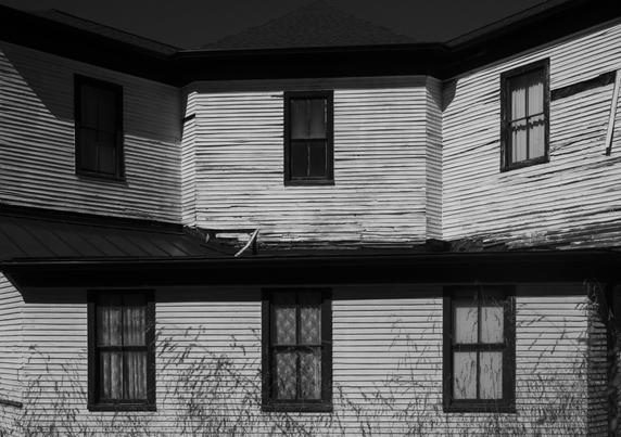 A high-contrast, black and white photograph of the side of an octagonal shaped two-story wooden building. The building features light-colored, peeling clapboard siding and dark trim around the windows. In the six windows are various curtain styles, including lace, horizontal blinds, fabric draperies, and some windows featuring no curtains at all. A dark, low-pitched roof separates the upper and lower floors, and dried, tall grass obscures the bottom of the first floor. The building's second story dark roof is set against an almost black sky.