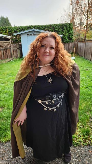 Me, a short, white pale woman with long curly red hair. I am wearing a brown wool cloak with a warm green linen lining. You can just see the inside of the hood draped over my back. I am wearing a long black dress with a lace corset belt and a silver chain belt that has various charms hanging from it like suns, moons, and hearts. I am also wearing a large silver mushroom necklace as well as a smaller silver necklace. The pose is giving Hobbit and the clothes are giving witch.