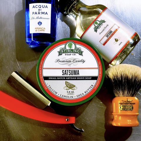 (Le Chat Mistral description)

This image shows a collection of traditional wet-shaving items arranged on a metallic surface.

At the top left, there is a blue glass bottle of **"Acqua di Parma Blu Mediterraneo Arancia di Capri"**.

In the centre, there is a round tin of **"Stirling Soap Co. Satsuma Small Batch Artisan Shave Soap"**, weighing approximately 5.8 ounces and made with tallow, lanolin, and shea butter.

To the right of the tin, there is a bottle of **"Stirling Satsuma Aftershave Splash"**.

At the bottom left, there is a straight razor with orange handles, partially unfolded.

At the bottom right, there is a yellow-handled **"Simpson Chubby 2 Manchurian Badger"** shaving brush.