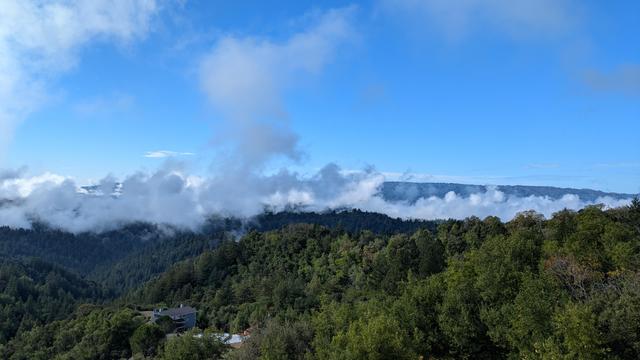 Looking down and across from a high ridge at puffy post-rain clouds of fog filling a valley in the middle distance, spilling over a low ridge into a partially sunny valley in the near foreground. The near valley is full of trees - redwoods and oaks - with a house visible in the lower part of the photo, just left of center. In the distance beyond the fog is a ridge, green with trees, and beyond that a barely visible line of ocean fog. The sky is bright blue and mostly clear, with a few small clouds in the left foreground, very close and about the same height as you are.