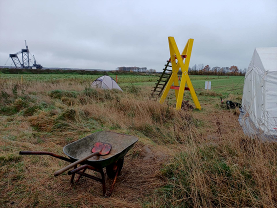 Hinter dem Mahnwachen-Zelt auf der Wiese des BUND steht ein großes, gelbes Holz-X, daneben ein Igluzelt. ImVordergrund eine Schubkarre mit Schaufel und im Hintergrund ein Schaufelradbagger im Tagebau Hambach, sowie am Horizont des "Sündenwädchen"