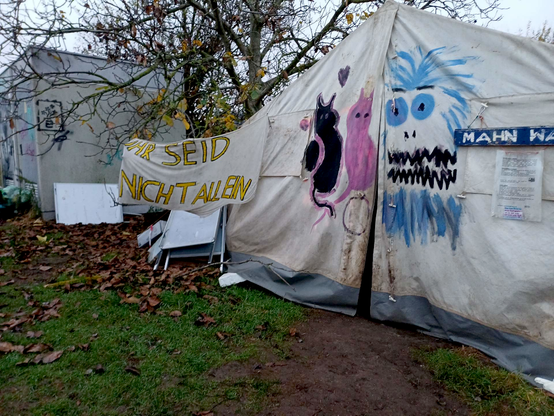 Ein weißes Gemeinschaftszelt steht an der Mahnwache Esperantsotraße und ist mit bunten Firguren bemalt. Daranhängt ein Banner: "Ihr seid nicht allein!", im Hintergrund ein kahler Laubbaum und ein Baucontainer.