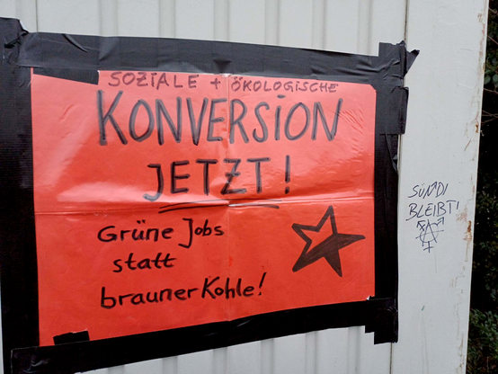 An einem Baucontainer bei der Mahnwache Esperantostraße hängt ein rotes Plakat: "Sozial-ökologisch Konversion jetzt! Grüne Jobs statt brauner Kohle!", darauf ein gezeichneter rot-schwarzer Stern. Daneben steht mit Filzstift geschrieben: "Sündi bleibt!"