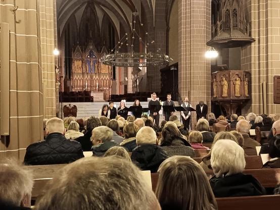 Blick durch ein Kirchenschiff zum Altar über die Köpfe vieler BesucherInnen hinweg. Vor dem Altar stehen die SängerInnen des Voktett Hannover unter einem großen Radleuchter.  Rechts daneben eine neugotische Kanzel, im Hintergrund ein neugotischer Hochaltar.
