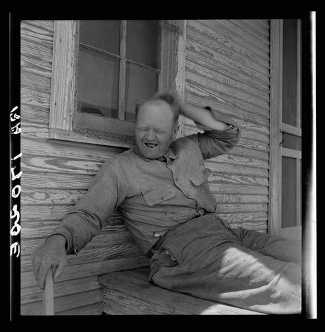 The black and white photograph captures a moment of levity or amusement on the porch of what appears to be an old wooden cabin, possibly indicative of rural life. The individual is seated comfortably with legs stretched out along the bench, one hand resting against their cheek while the other hovers in mid-motion as if brushing back hair from their face. They wear a sleeveless shirt and shorts typical for warmer weather or activity-oriented workwear, suggesting practicality over formality. Beside them on the ground is what seems to be an old-fashioned cane with a worn grip, hinting at mobility concerns.

The setting of this photograph carries historical significance, being part of Dorothea Lange's series documenting sharecroppers in Texas during the Great Depression era as detailed by "Sharecropper" and identified further via metadata reference. This context adds layers to interpreting the image; it is not just a casual snapshot but an important social document shedding light on life conditions at that time.

The photograph carries a certain graininess characteristic of film from its period, emphasizing texture over clarity which contributes to evoking nostalgia or historicity. Despite being in grayscale, textures are well-defined — the wood's worn look and the person’s clothing all hinting toward an era long past but captured with enduring relevance through Lange's lens.

This image is a part of Dorothea Lange's extensi [...]