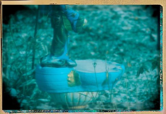 A plastic camera setting gives the composition a soft focus & blue tone, with darkened vignette. A birdbath on a pedestal is filled with ice, and a vert light dusting of snow clings to the grass and plants in the background.