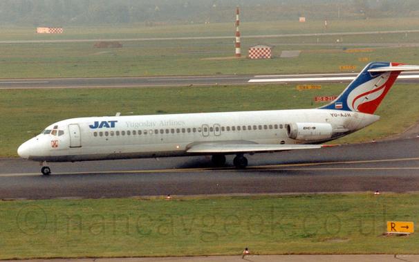 Side view of a twin engined jet airliner with the engines mounted on the sides of the rear fuselage, taxiing from right to left along a black taxiway.
The plane is mostly white, with a slivery belly, and blue "JAT" titles  on the upper forward fuselage, followed by grey "Yugoslav Airlines" text.
There is black "DC-9" text on the white engine pods, and the black registration "YU-AJH" on the upper rear fuselage, under the tail.
The tail is blue at the front and red at the rear, overlaid with a stylised white flame.
Green grass in the foreground lines the taxiway, as well as runways in the background, , with trees in the distance, slowly vanishing in to haze.