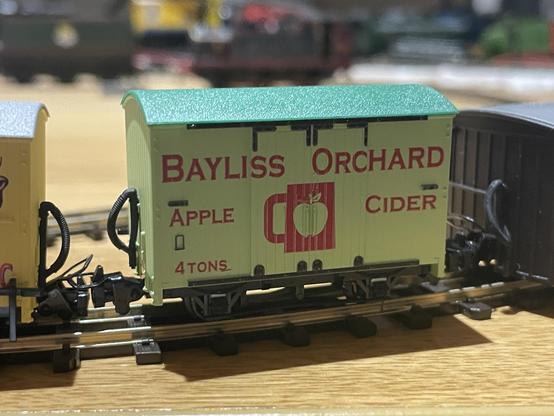 A small model railway van in a pale green colour with “Bayliss Orchard Apple Cider” written on the side in red. In the middle is a mug shape with an apple in it