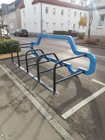 Auf einem ehemaligen Parkplatz steht jeutzt j Fahrradständer mit 4 Buchten. 10 Räder könknten hier angeschlossen werden. Diese stabile Konstruktion erinnert der  Form nach an 
ein Auto