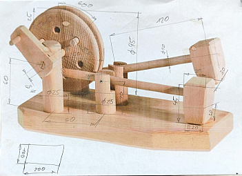 Holzspielzeug-Modell einer kleinen mechanischen Maschine: Auf einer rechteckigen Holzplatte sind mehrere Klötze, Hebel und ein großes rundes Schwungrad montiert. Darüber liegen handgezeichnete Maßangaben, die verschiedene Teile des Spielzeugs markieren.