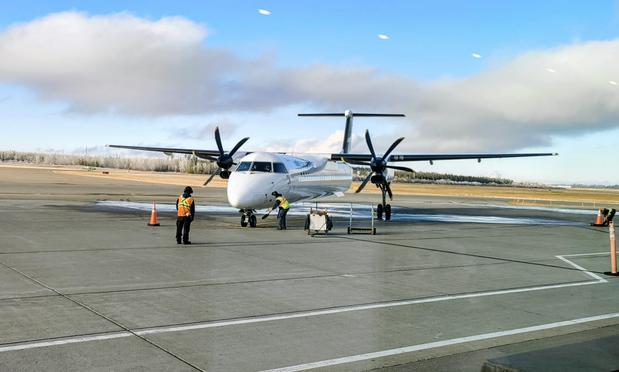 Showing a high wing turboprop passenger aircraft with a high T tail configuration. The propellers are six bladed each and there are two of them. The landing gear extends from the engine nacells on each side. 

There are ground personel guiding it in and someone putting wheel blocks on the nosewheel.

The plane is mostly white.

The sky is party cloudy with blue sky.