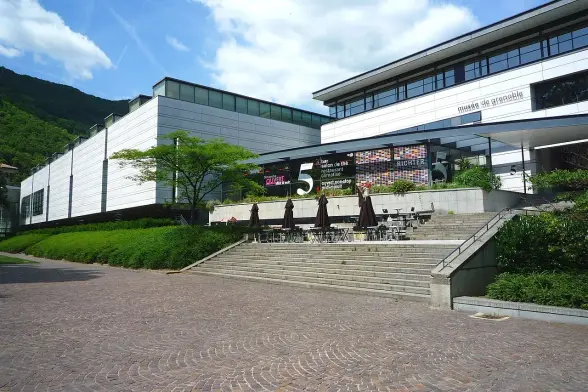The image features a modern building with a sleek architectural design, likely part of a cultural or educational institution. It has large windows and a prominent entrance. In the foreground, there are stairs leading up to a terrace with outdoor seating under umbrellas. The surrounding area includes greenery, enhancing the outdoor atmosphere. The building is set against a backdrop of hills or mountains, suggesting a picturesque location. The signage indicates the presence of an exhibit or event.

Image Credits: Wikimedia / Milky / CC BY-SA 3.0