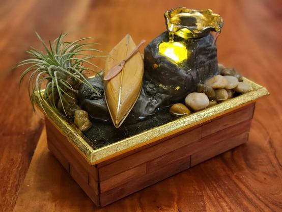 The fountain sports warm wooden siding, shining gold accents, and a real air plant. It has been painted and textured to look like rocks, and a glossy miniature cedar-strip canoe sits upside down on the rocks. A paddle lies across the canoe's keel.