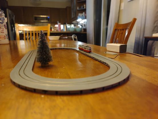 A red tram the size of an adult human finger runs around an oval of streetcar track on a wooden table, surrounding a single miniature pine tree speckled with fake snow.