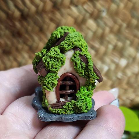 I am holding a very tiny polymer clay house, maybe 2 inches tall and an inch wide. It has tan walls, a dark wooden roof that is covered in green moss. And then a small window in a shiny copper. It is sitting on a small piece of speckled grey clay.