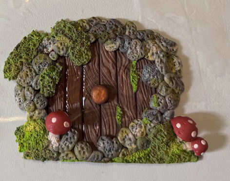 A round door with a wood effect, surrounded by rocks and moss. It has a copper knob in the centre and three small mushrooms around it.