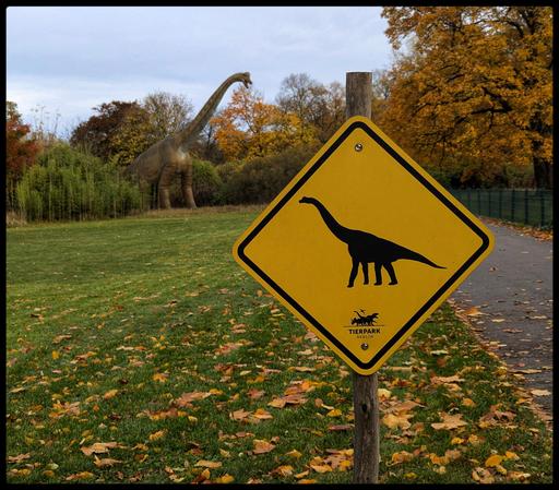 Im Vordergrund ein gelbes Hinweisschild, dass Dinosaurier kreuzen können. Im Hintergrund kommt ein Dinosaurier aus dem Gebüsch hervor.